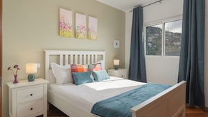 Un dormitorio con una cama blanca, una mesita de noche con lámpara y una ventana con cortinas.