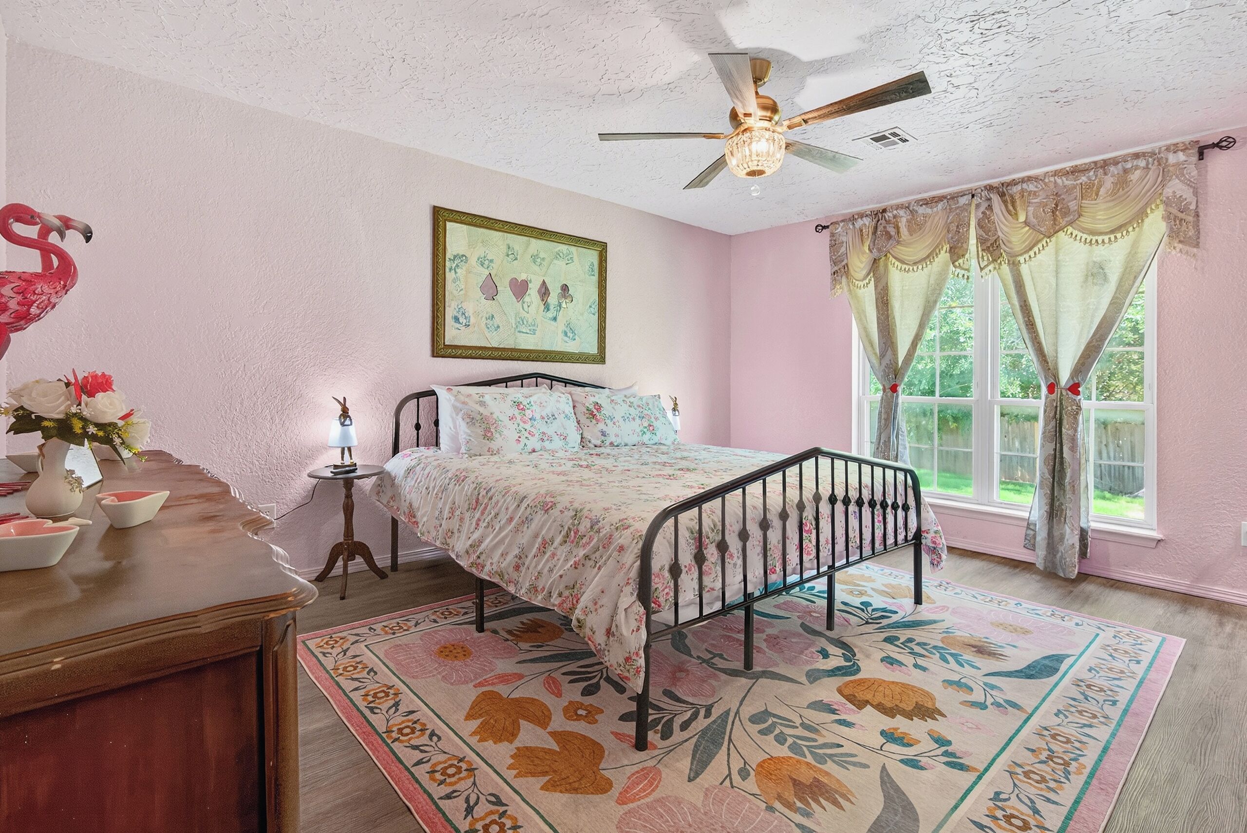 Une chambre à coucher avec un lit, un ventilateur de plafond, une fenêtre avec des rideaux, une table de chevet avec une lampe et un tapis à motifs floraux.