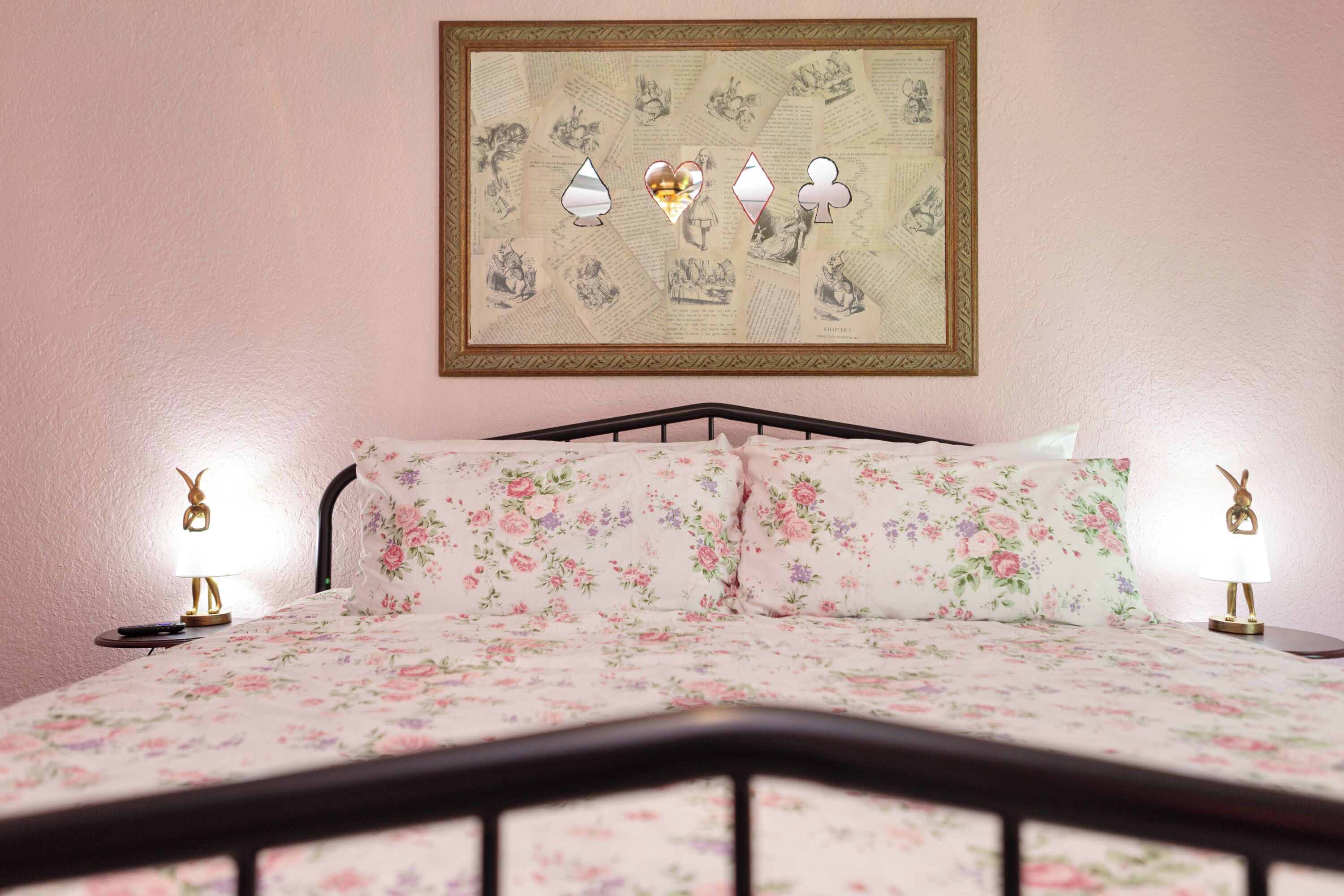 A bed with floral bedding, two bedside lamps with animal-shaped bases, and a framed picture with playing cards on the wall.