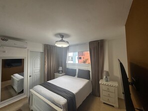 A bedroom with a bed, bedside tables, a mirror, a TV, and an air conditioning unit.