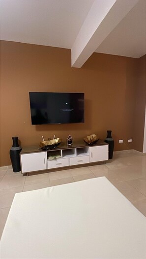 A living room with a flat-screen TV mounted on the wall, a white entertainment unit with decorative items, and a large white rug on the floor.