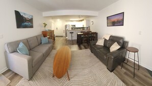Ein modernes Wohnzimmer mit Sofa, Couchtisch und einem Essbereich mit Stühlen, im Hintergrund eine Küche.