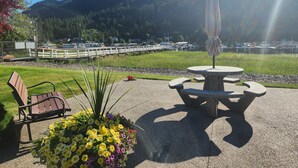 Eine Terrasse mit Tisch und Stühlen, einem Blumenbeet und Blick auf einen Steg sowie Boote auf einem See, im Hintergrund Berge.