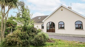 A single-story house with a sloped roof, large windows, and a well-maintained garden with various plants and a palm tree. - LILLY'S BUNGALOW, pet friendly in Clonakilty, County Cork (Timoleague)