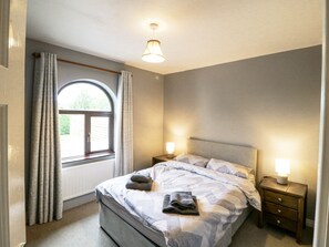 A bedroom with a bed, two bedside tables with lamps, a window with curtains, and a radiator. - LILLY'S BUNGALOW, pet friendly in Clonakilty, County Cork (Timoleague)