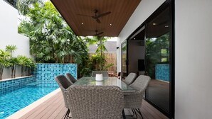 Un patio moderno al aire libre con mesa de vidrio, sillas de mimbre, piscina y abundante vegetación.