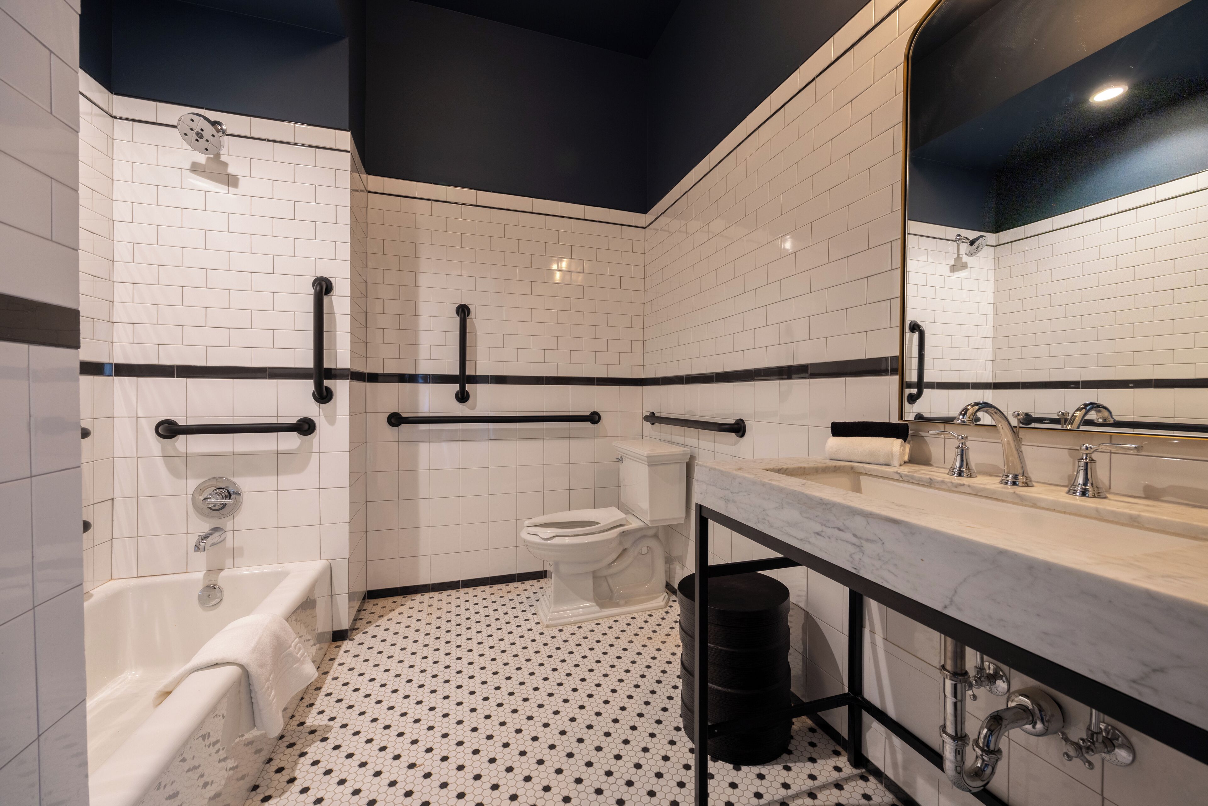 A modern bathroom with white subway tiles, a black and white patterned floor, a large mirror, a bathtub, a toilet, and a sink with a marble countertop.