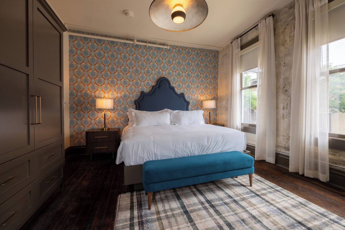 Un dormitorio con una cama grande, dos lámparas de noche, un banco azul, un papel tapiz estampado y una ventana con cortinas.