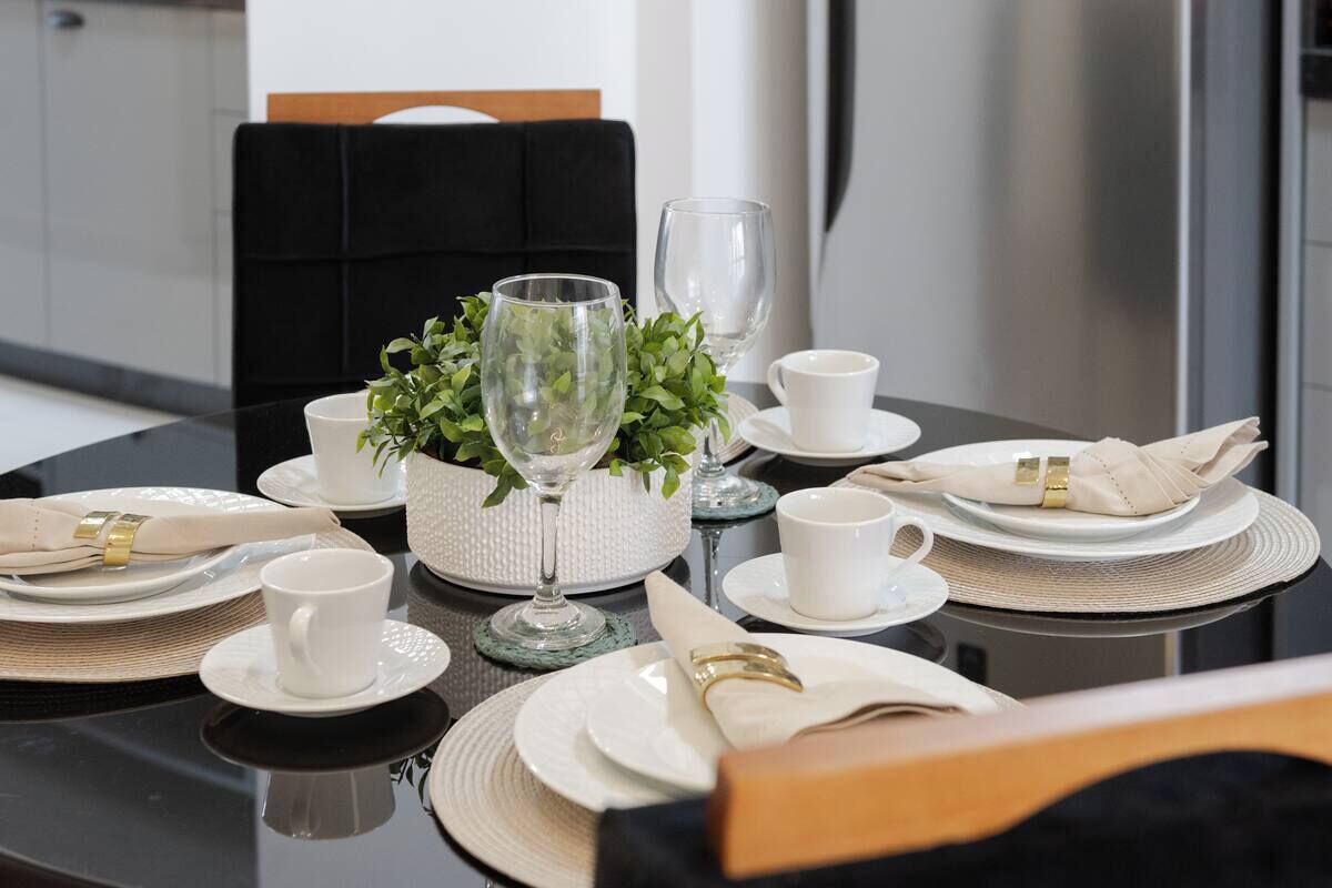 A dining table set with white plates, bowls, and cups, accompanied by a decorative centerpiece of green foliage and a glass.