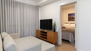 Un salon moderne avec un canapé, un téléviseur posé sur un meuble en bois et une porte donnant sur une chambre à coucher équipée d’un lit et ornée d’un tableau.