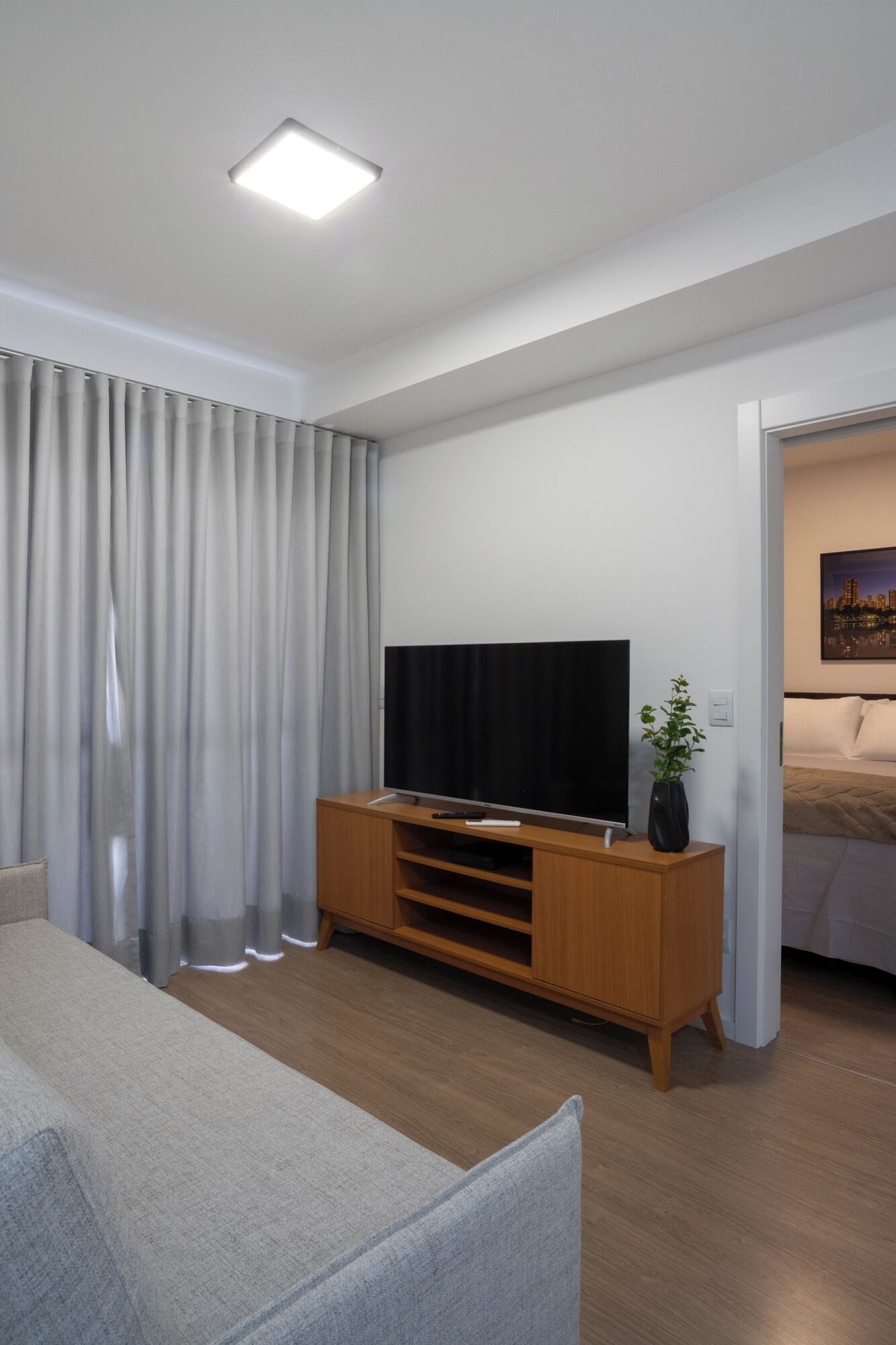 Ett modernt vardagsrum med en platt-TV placerad på ett träställ, en grå soffa och ett stort fönster med grå gardiner.