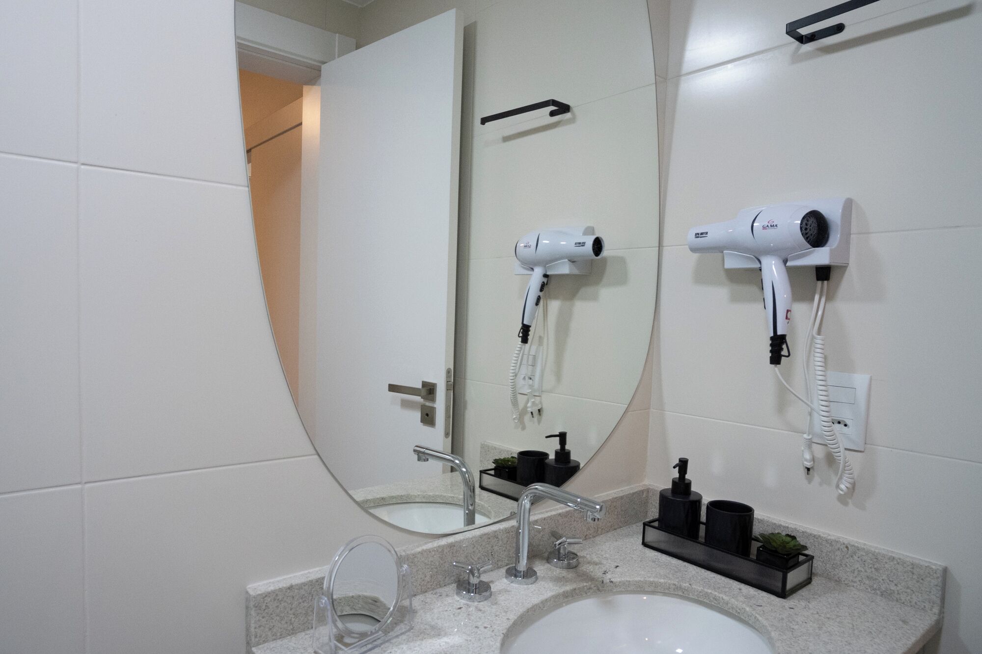 A bathroom with a large mirror, two hair dryers mounted on the wall, a sink with a faucet, and a small shelf with toiletries.