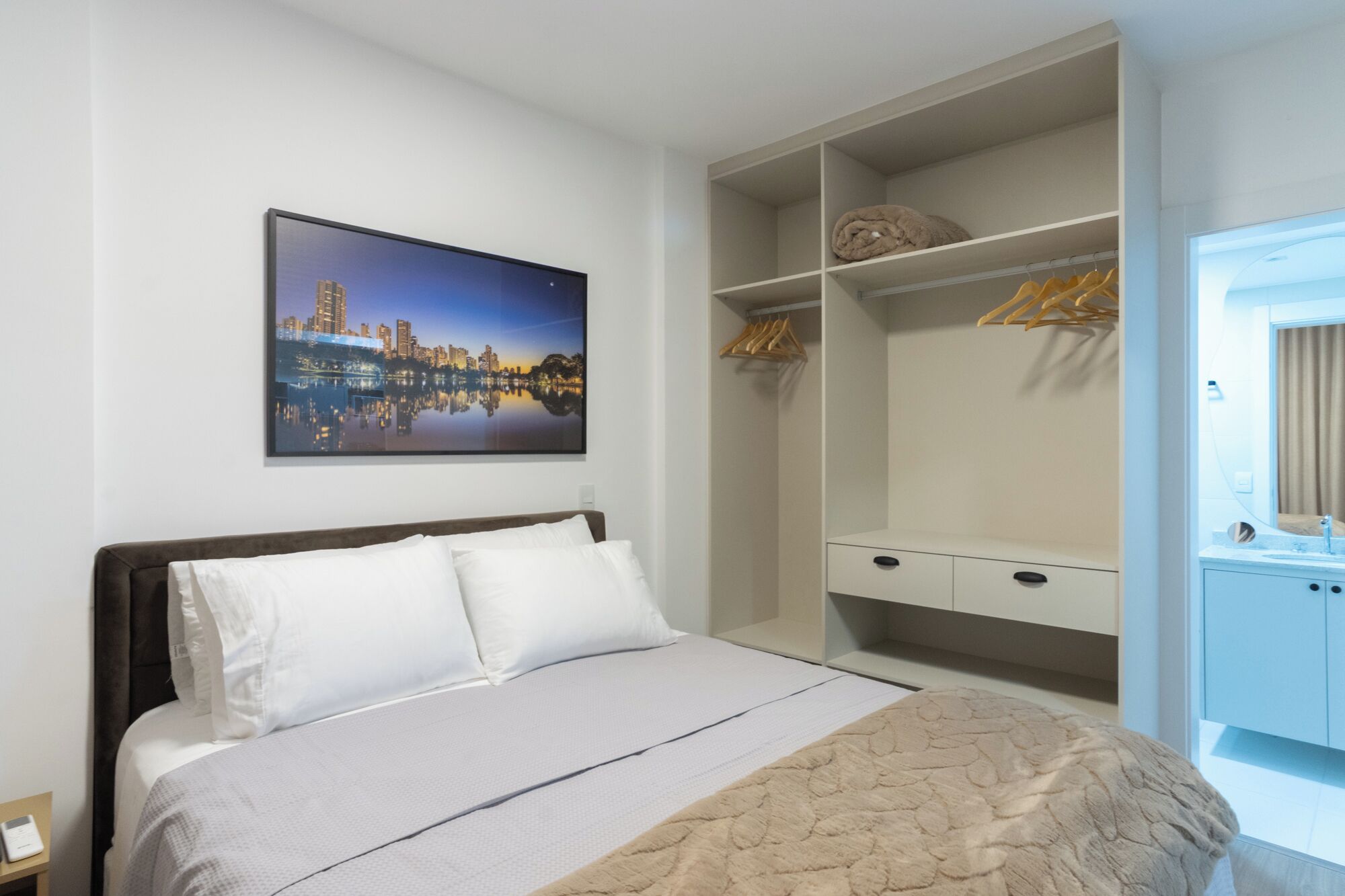 A hotel room with a bed, a wardrobe with hanging space, and a cityscape painting on the wall.