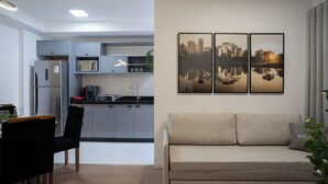 Une cuisine moderne avec des meubles gris, un réfrigérateur en acier inoxydable et un four à micro-ondes. On y trouve un coin repas avec une chaise noire et une table. Un canapé beige se trouve dans le salon, au-dessus duquel est accrochée une peinture représentant un paysage urbain.