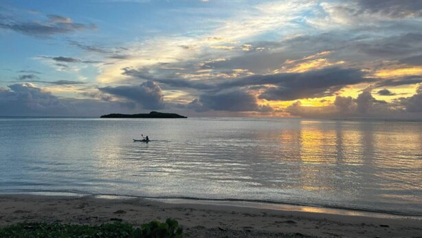 A beach with calm waters, a distant island, and a clear sky with clouds.