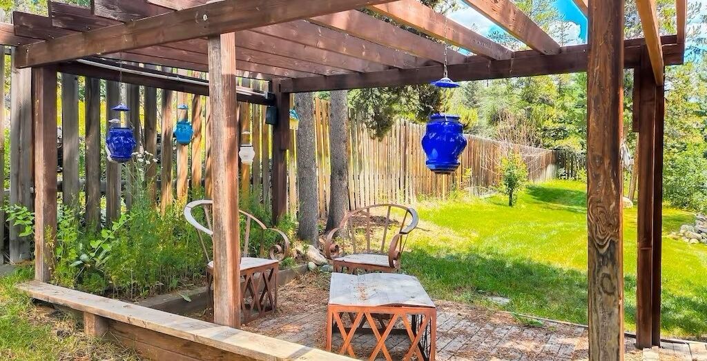 Een houten pergola met hangende blauwe lantaarns, een houten bank en een tafel met stoelen in een tuin.