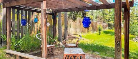 Een houten pergola met hangende blauwe lantaarns, een houten bank en een tafel met stoelen in een tuin.