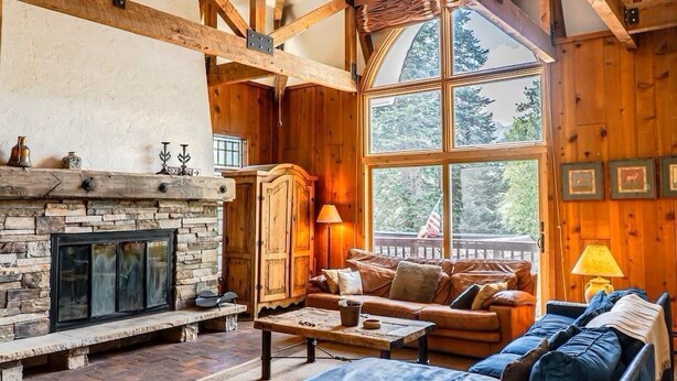 A cozy log cabin interior with a stone fireplace, wooden beams, a large window, and a comfortable seating area. - Lodge Off Main, Walk to Gondola! (Breckenridge)