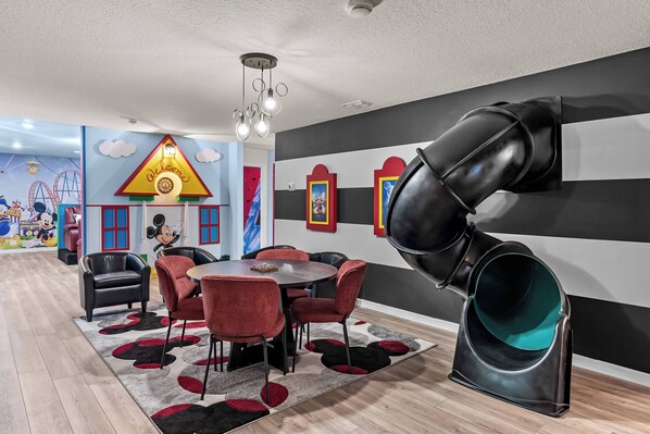 A children's play area with a slide, a round table with chairs, and a mural with cartoon characters.