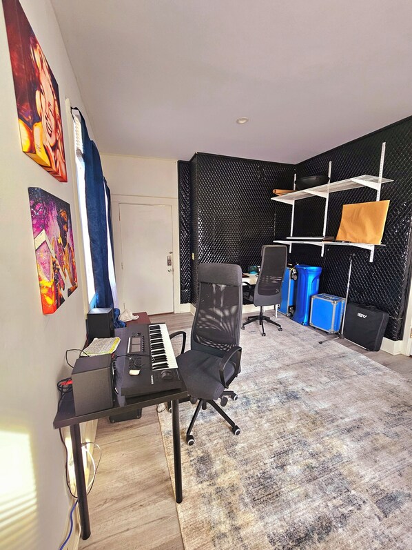 A room with a desk, a chair, a keyboard, and a computer. There are two paintings on the wall, a window with curtains, and a black acoustic panel on the wall.