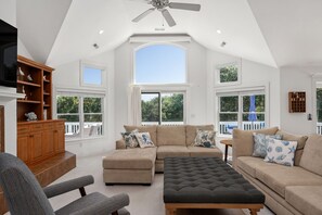 A spacious living room with a high ceiling, large windows, and a ceiling fan.