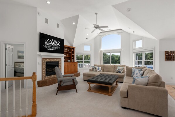 A spacious living room with a fireplace, a television mounted on the wall, and a large window providing natural light.