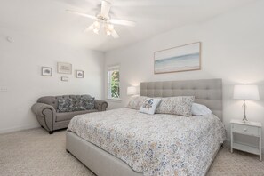 Ein Schlafzimmer mit einem großen Bett, einem grauen Sofa, einem Deckenventilator und einem Fenster mit Blick auf Grünflächen.