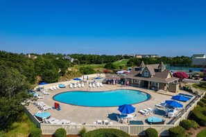 A large swimming pool with lounge chairs and umbrellas, surrounded by a wooden deck and a building with a pitched roof.