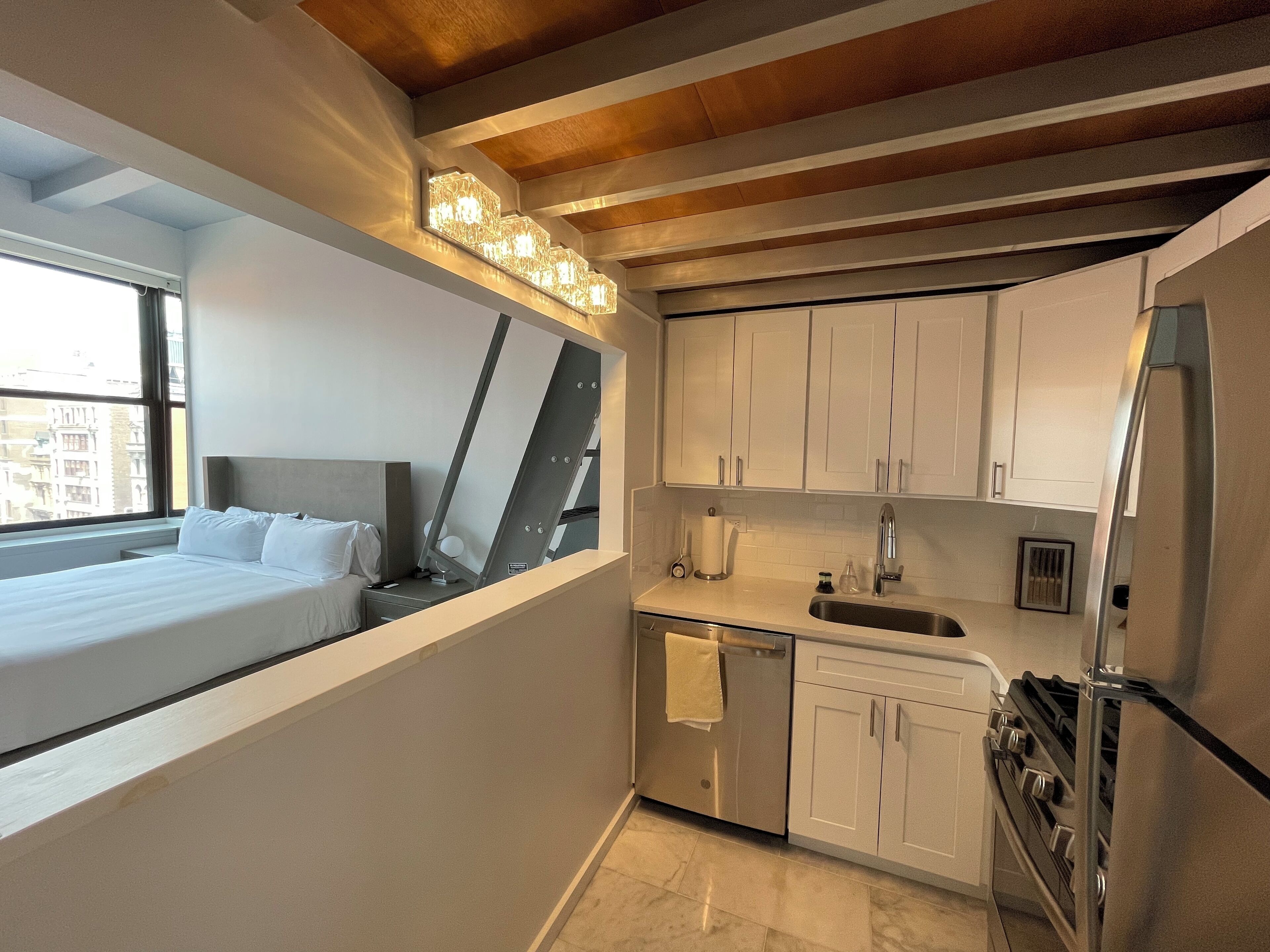 A modern hotel room with a bed, a large window, a kitchen with white cabinets, a stainless steel refrigerator, and a built-in oven.