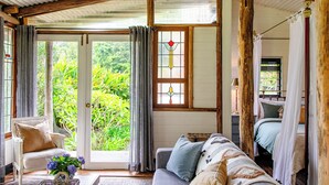 A cozy room with a sofa, a chair, and a bed. There is a window with curtains and a view of greenery outside.