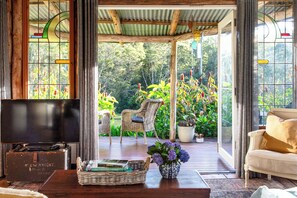 A cozy living area with a television, wicker chairs, a sofa, and a coffee table with books and a vase of flowers. - Heritage Cabin | Clawfoot Bath in the Rainforest | Byron Bay Hinterland (Dorroughby)
