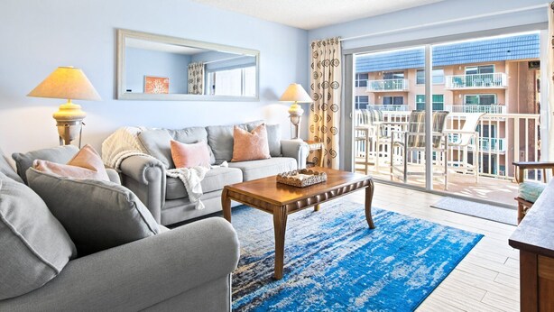 A living room with a grey sofa, a wooden coffee table, and a large window with a view of a balcony and building. - Cocoa Beach Oceanfront | Sunrise Views, Heated Pool & BBQ Grill | Walk to Downtown (Cocoa Beach)