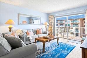 Een woonkamer met een grijze bank, een houten salontafel en een groot raam met uitzicht op een balkon en een gebouw.