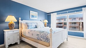 A bedroom with a bed, two nightstands, a window with a view of buildings, and a picture on the wall. - Cocoa Beach Oceanfront | Sunrise Views, Heated Pool & BBQ Grill | Walk to Downtown (Cocoa Beach)