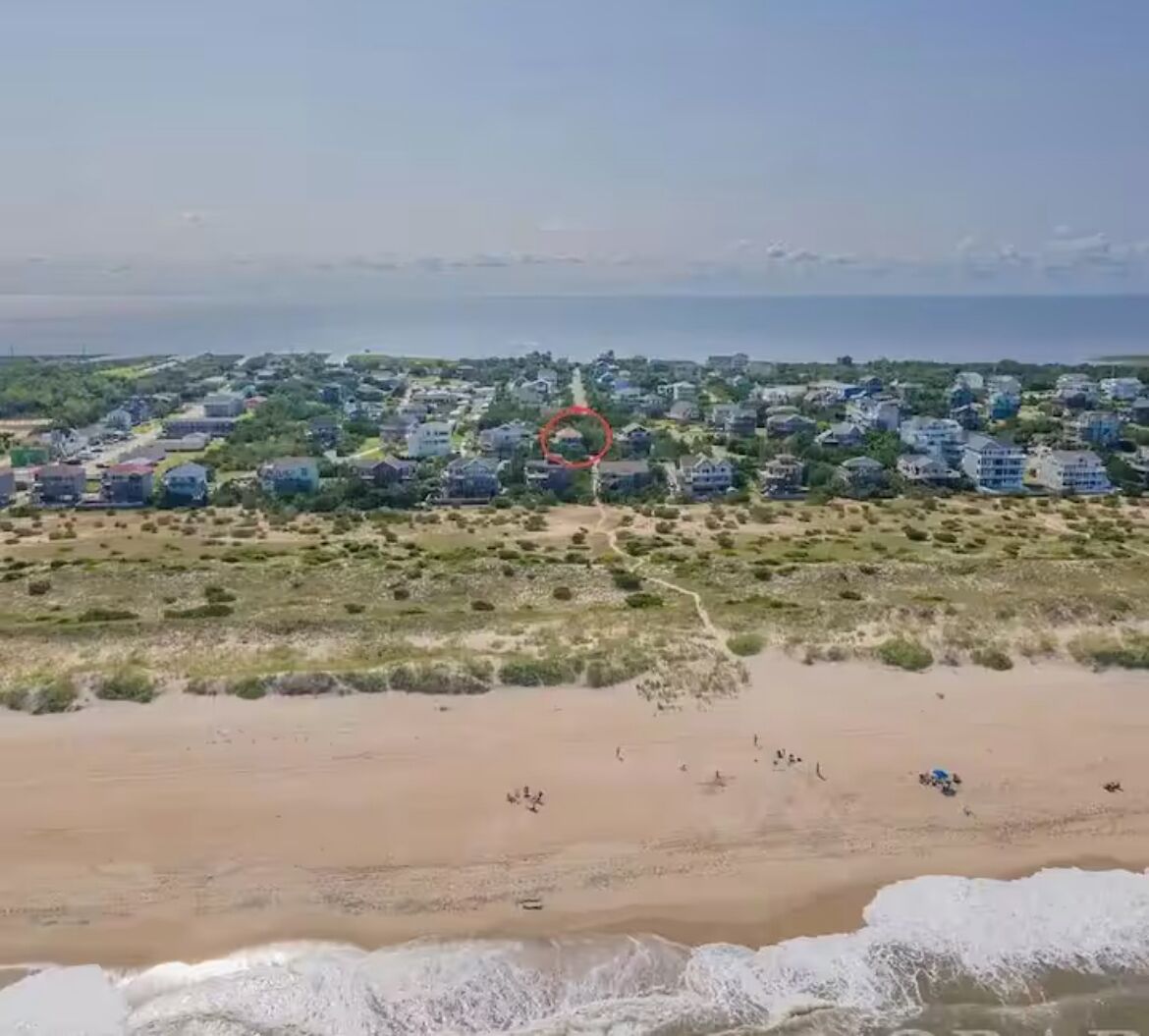 An aerial view of a coastal area with a sandy beach, a path leading to a cluster of buildings, and a body of water in the background.