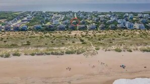 Vista aérea de una zona costera con playa arenosa, un camino que lleva a un conjunto de edificios y una masa de agua al fondo.