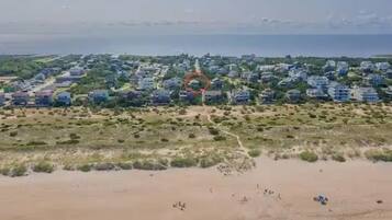 An aerial view of a coastal area with a sandy beach, a path leading to a cluster of buildings, and a body of water in the background.