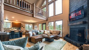 A spacious living area with a fireplace, a kitchen in the background, and a balcony with wooden railings. - Epic Views + Stargazing Net • Hot Tub • Sauna • Luxury Vibes (Fairplay)