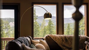A cozy room with a view of mountains and trees through large windows, a brown leather sofa, and a floor lamp. - Epic Views + Stargazing Net • Hot Tub • Sauna • Luxury Vibes (Fairplay)