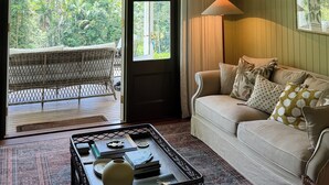 A cozy living room with a large window, a wicker chair, a sofa with cushions, a coffee table, and a hanging light fixture. - Rainforest Retreat | Fire, Stars & Birdsong | Byron Bay Hinterland (Dorroughby)