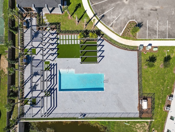 An aerial view of a parking lot with a swimming pool, a covered area, and a fenced section with a small building.