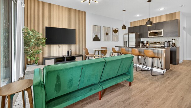 A modern living room with a green sofa, wooden panel walls, a flat-screen TV, and a kitchen area with bar stools. - 3231 ED - The Hub Modern 3BR (Kissimmee)