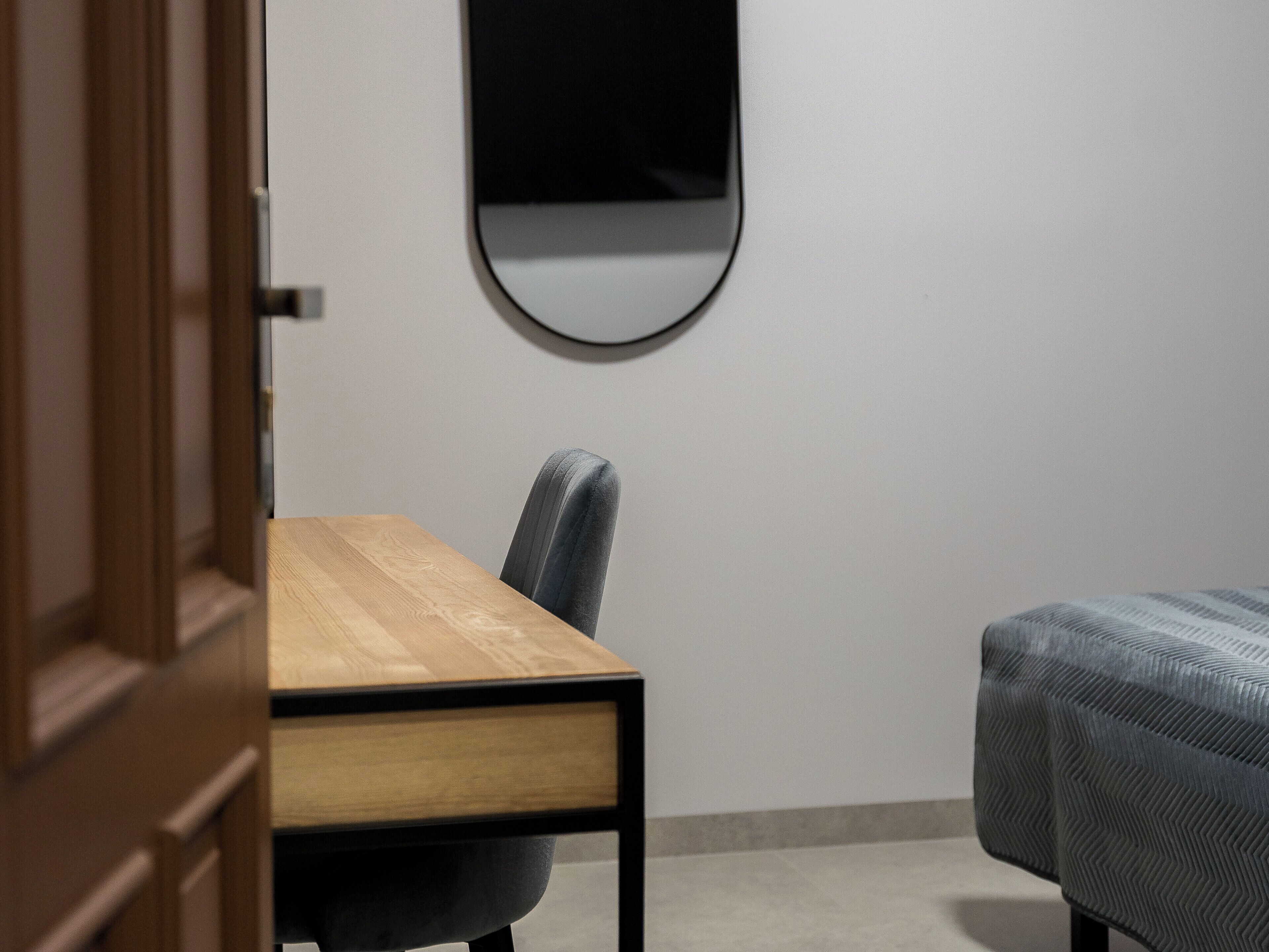 Une pièce avec un bureau en bois, une chaise, un lit recouvert d’une courtepointe à motifs et un miroir rond accroché au mur.