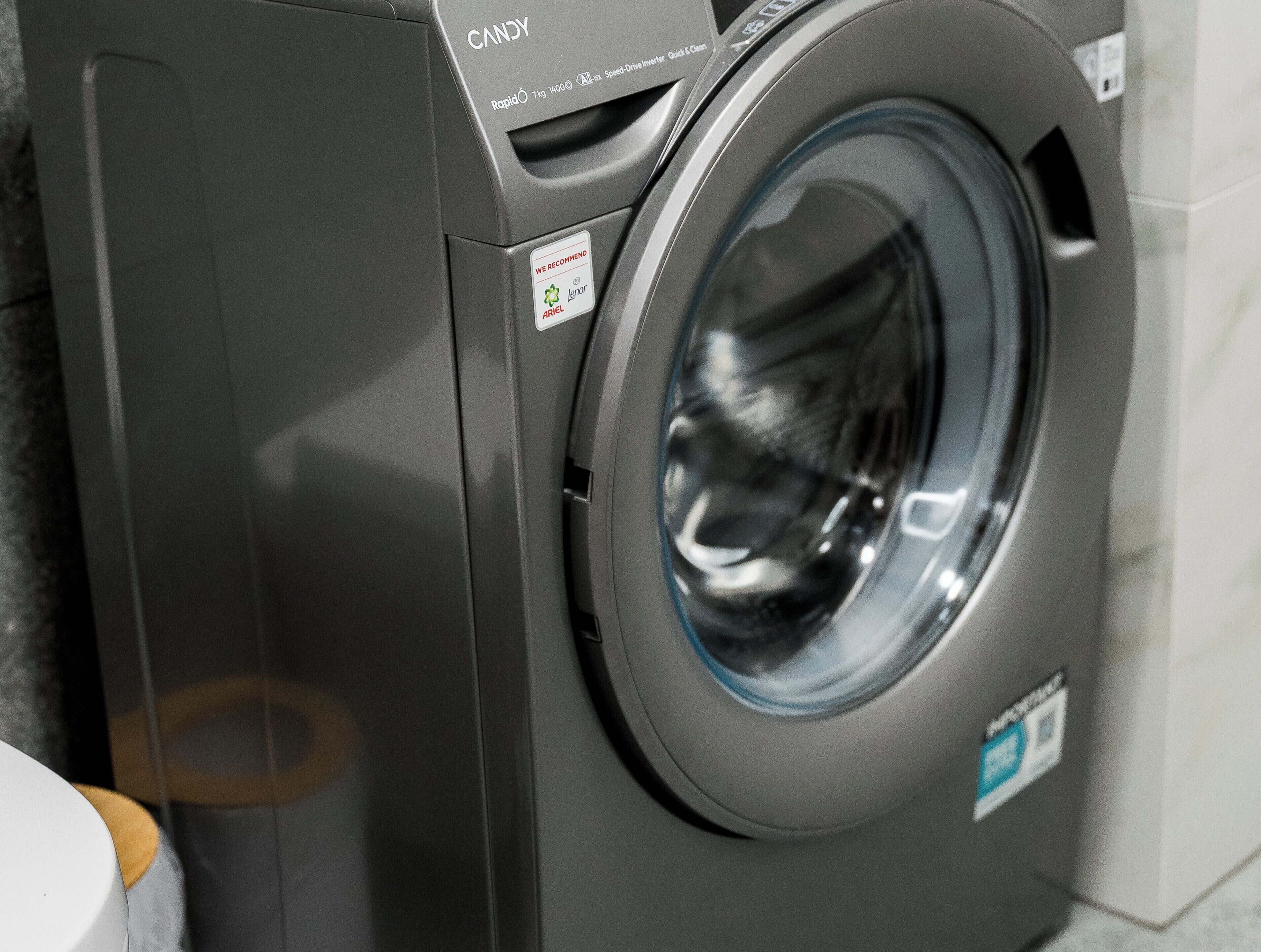 A front view of a front-loading washing machine with a control panel and a label recommending Ariel detergent.