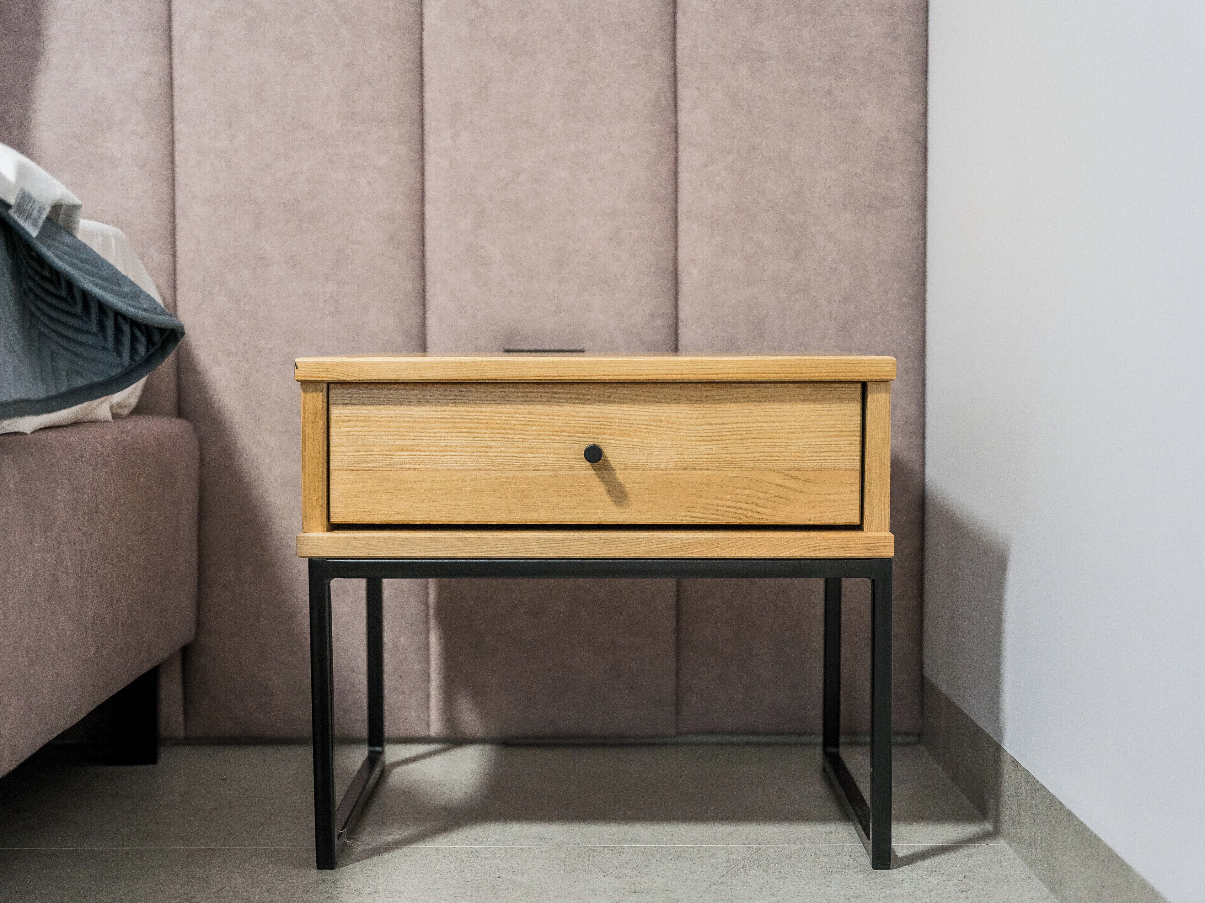 Un comodino in legno con un cassetto, gambe in metallo nero e un cuscino su un divano sullo sfondo.