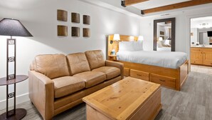 A modern hotel room with a wooden bed, a leather sofa, a wooden coffee table, and a small side table with a lamp. - Newpark 217 | Private Hot Tub | Kitchenette | Fireplace | Washer & Dryer | Balcony | Pool & Hot Tub (Park City)