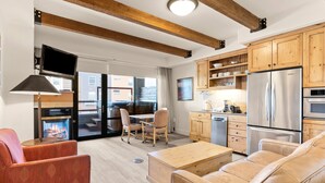 A modern living room with a sofa, a coffee table, and a fireplace. There is a kitchen area with wooden cabinets and a stainless steel refrigerator.