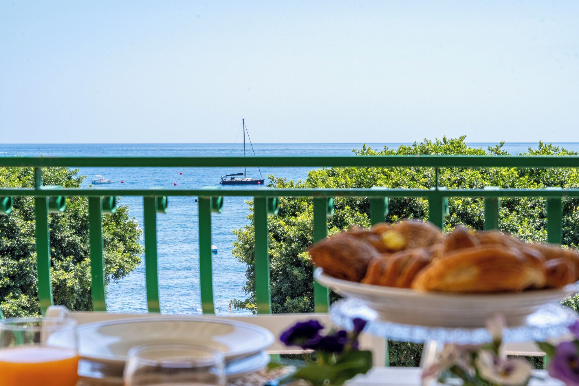 Un balcon avec vue sur la mer, une assiette de pâtisseries et un verre de jus d’orange.