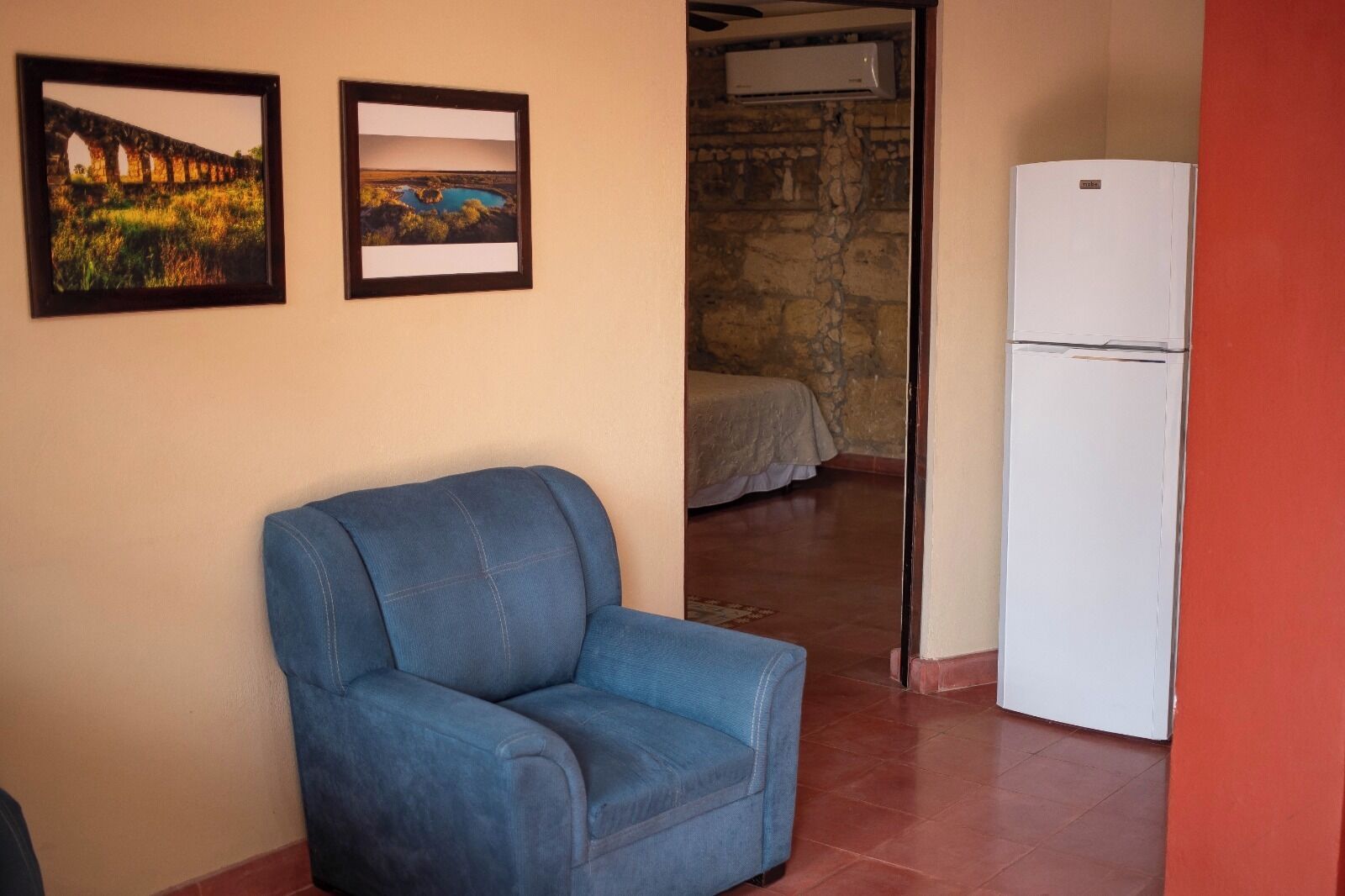 Een kamer met een blauwe fauteuil, een bed, een witte koelkast en twee foto's in lijsten aan de muur.