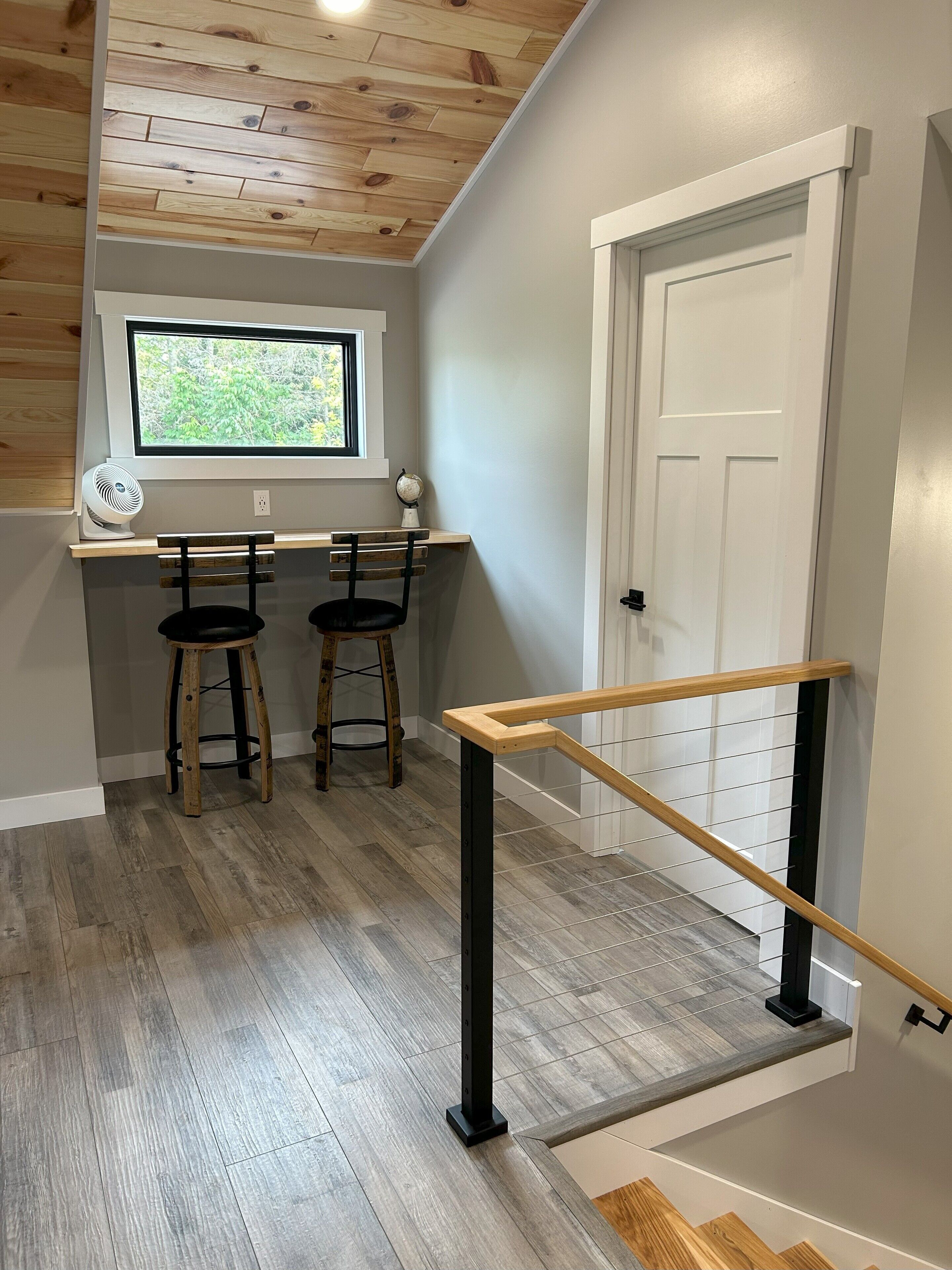 Una habitación con techo de madera, ventana con vistas, un escritorio y dos sillas, y una puerta blanca.
